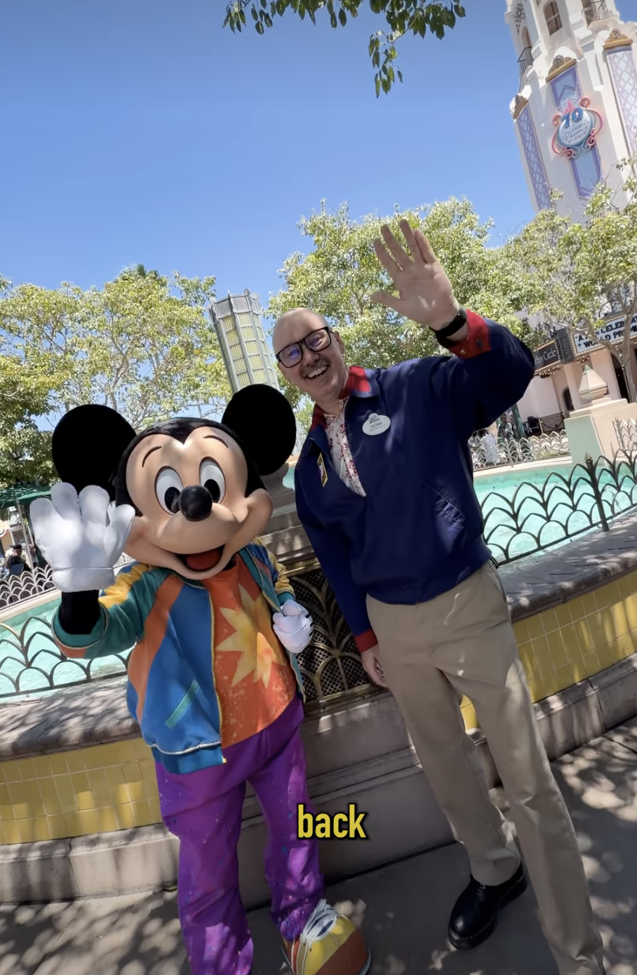 Mickey Mouse and a Cast Member stand beside a fountain and wave toward the camera