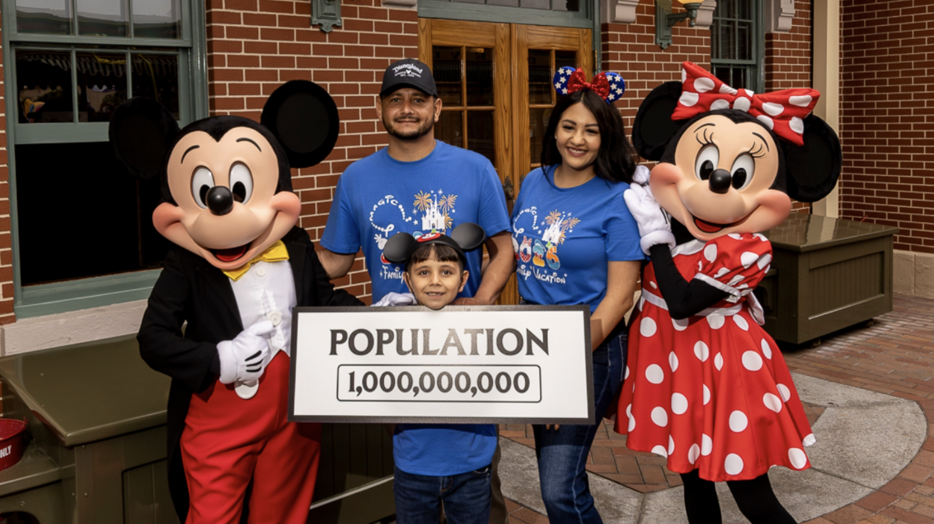 Mickey Mouse and Minnie Mouse stand with a family of three holding a sign that reads “Population 1,000,000,000” in front of a brick building at Disneyland Resort, celebrating the resort’s one billionth guest milestone.
