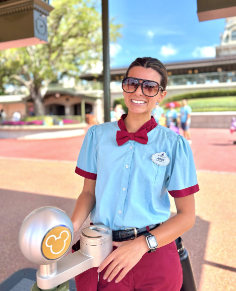 Cast Member in blue short sleeve button down with red bow tie smiling in front of Magic Kingdom ticket reader