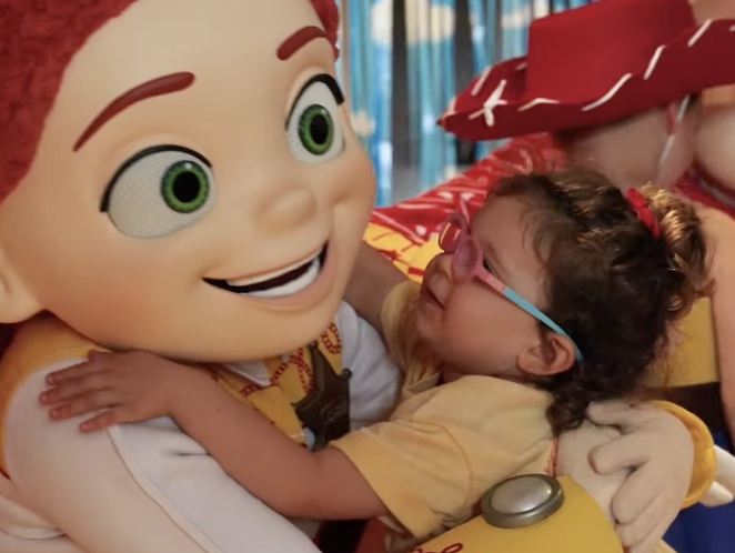 Jessie from Toy Story hugs a young child indoors while another character from Toy Story is partially visible in the background.