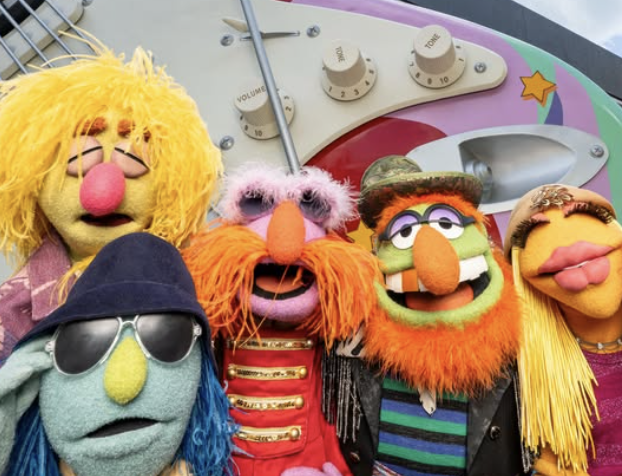 Several Muppets characters pose together outdoors in front of colorful theme park signage and decorations. The group is gathered closely for a photo with palm trees and a bright sky visible in the background.
