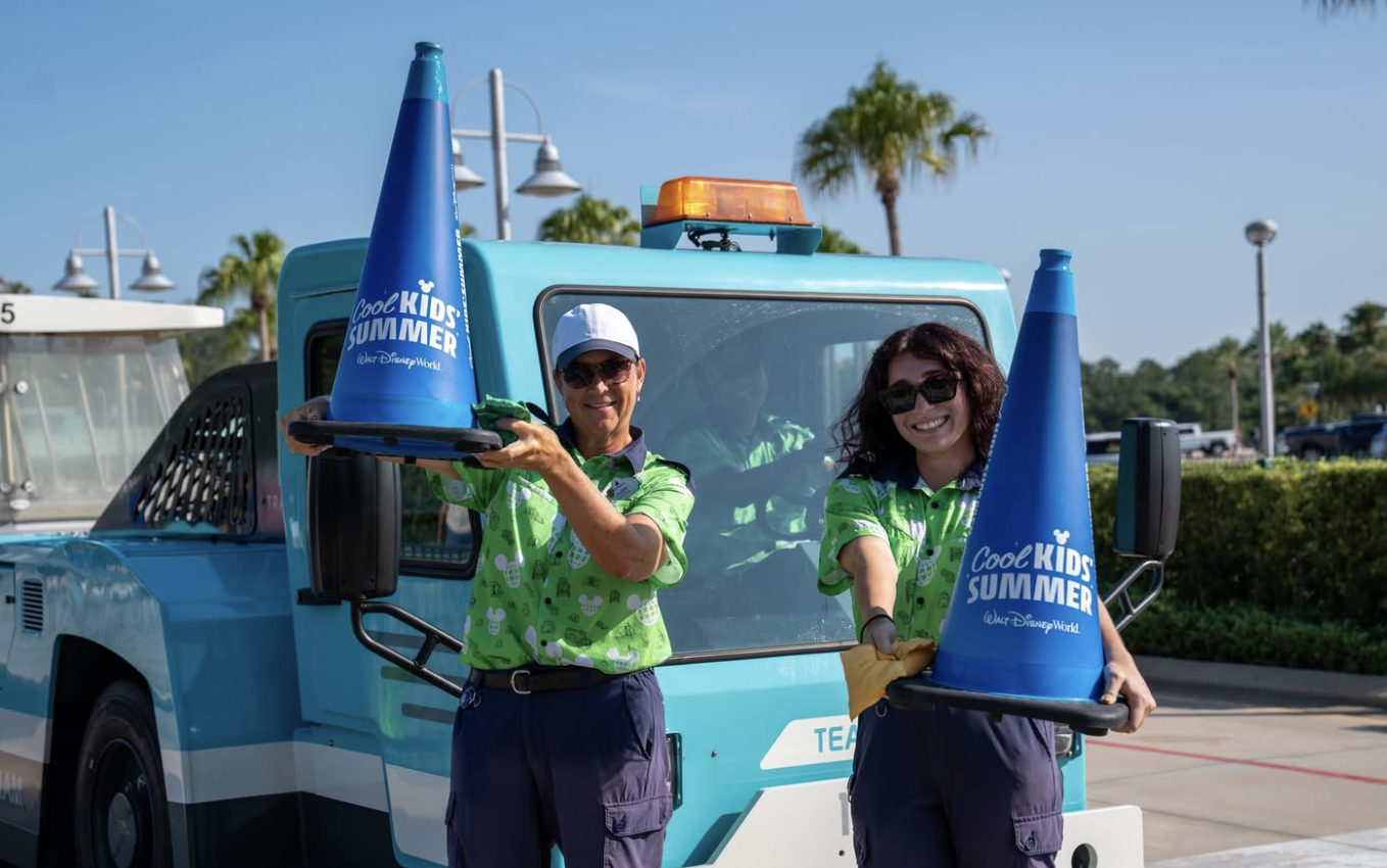 Two Walt Disney World Cast Members stand in front of a blue service vehicle holding oversized blue traffic cones branded with the “Cool Kid Summer” Walt Disney World logo. Palm trees and a sunny outdoor setting are visible in the background.