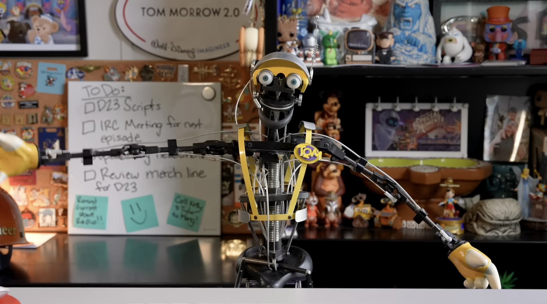 A robot character with yellow accents extends its arms in a workspace filled with Disney collectibles, sketches and memorabilia.