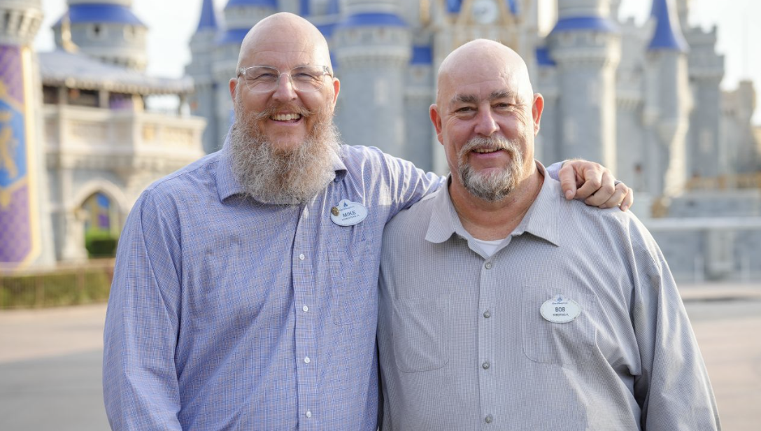 Two Walt Disney World Cast Members stand arm in arm in front of Cinderella Castle, posing together for a photo.