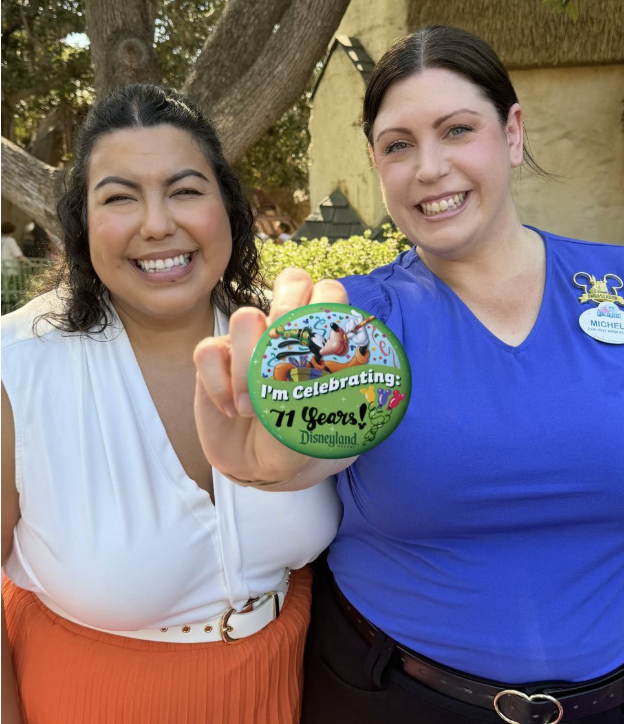 Two Disneyland Resort Cast Members hold a commemorative button celebrating the resort's 71st anniversary toward the camera while standing outdoors.