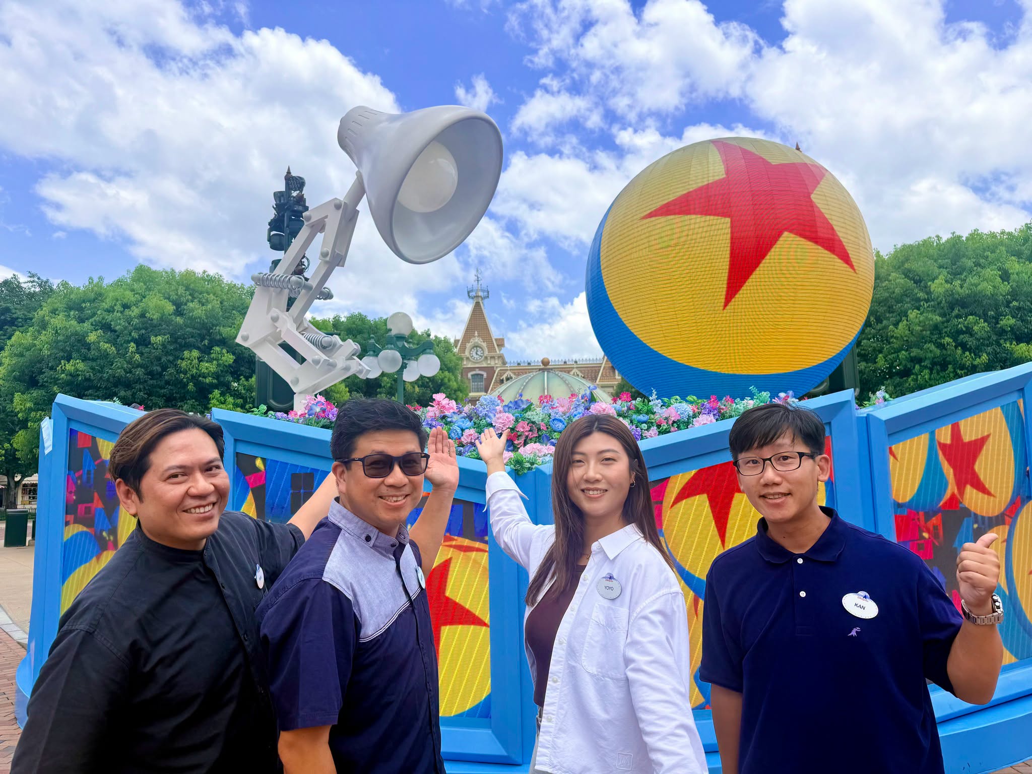 Four Hong Kong Disneyland Cast Members stand in front of a large Pixar Luxo Jr. lamp and Pixar ball displa